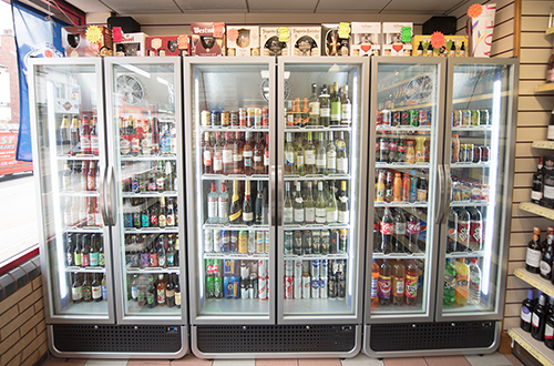 Four glass-door refrigerators display various beverages, including soda cans, wine bottles, and beer, in a brightly lit convenience store. Shelves around the fridges hold additional drink options.