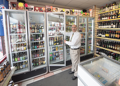 A man in formal attire opens a glass door refrigerator stocked with various beverages in a well-lit convenience store. Shelves filled with bottles are on either side, with a freezer in the foreground.