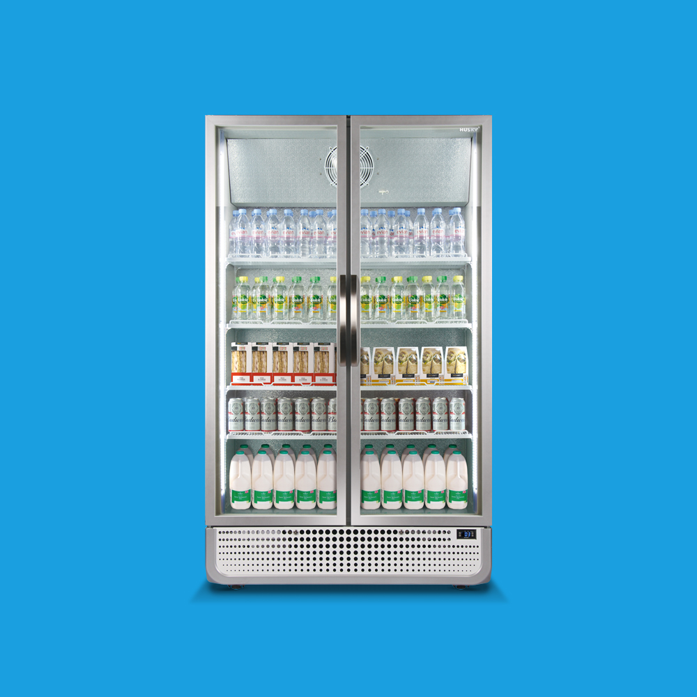 A glass-door refrigerator filled with bottled water, cans, and various dairy products stands against a bright blue background. The fridge is well-organized, showcasing items evenly spread across five shelves.