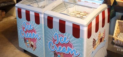 Large ice cream freezers, adorned with "Ice Cream" and "Frozen Treats For Everyone!" signs, sit side by side in a store selling various food items, including fresh produce and baked goods.