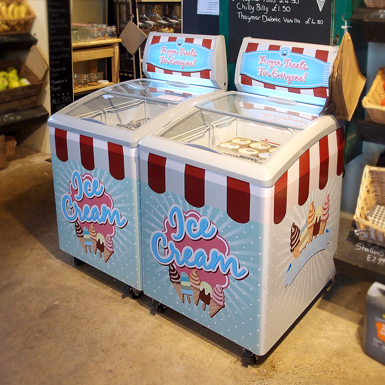 Large ice cream freezers, adorned with "Ice Cream" and "Frozen Treats For Everyone!" signs, sit side by side in a store selling various food items, including fresh produce and baked goods.