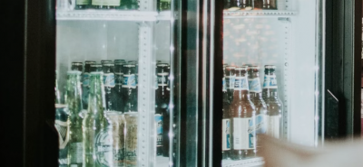Bottles of beer sit inside a well-lit refrigerated display case, with various brands visible; the display case is located within what seems to be a bar or store.