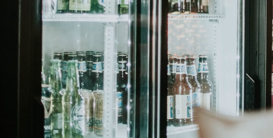 Bottles of beer sit inside a well-lit refrigerated display case, with various brands visible; the display case is located within what seems to be a bar or store.