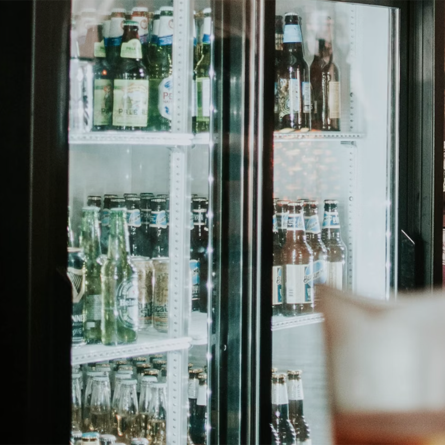 Bottles of beer sit inside a well-lit refrigerated display case, with various brands visible; the display case is located within what seems to be a bar or store.