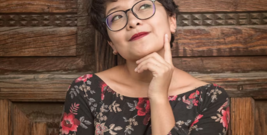 Person wearing glasses and a floral dress, thoughtfully resting their chin on their hand, standing against a rustic wooden backdrop with a decorative pattern.