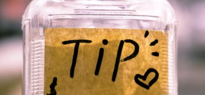A clear jar labeled "TIP" with money inside sits on a counter in a blurred background, suggesting a cafe or store environment.