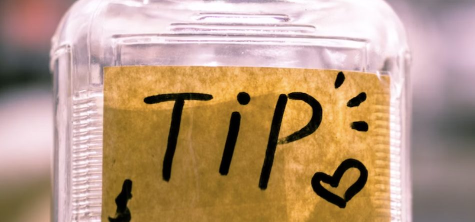 A clear jar labeled "TIP" with money inside sits on a counter in a blurred background, suggesting a cafe or store environment.