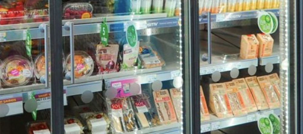 A refrigerated supermarket display case contains prepared salads, sandwiches, drinks, and packaged meals, all organized on shelves with price tags and green promotional signage visible.