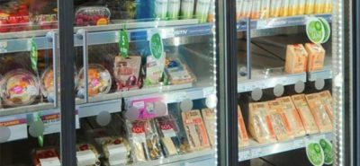 A refrigerated supermarket display case contains prepared salads, sandwiches, drinks, and packaged meals, all organized on shelves with price tags and green promotional signage visible.
