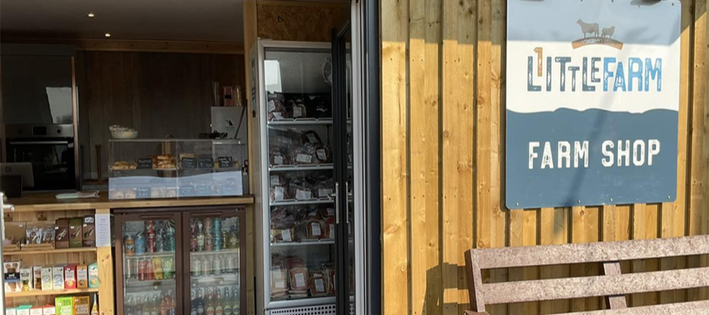 Store counter displays various goods; behind glass are baked items, below are beverages. The setting is a wooden farm shop with a sign reading “LITTLE FARM FARM SHOP” near a bench outside.