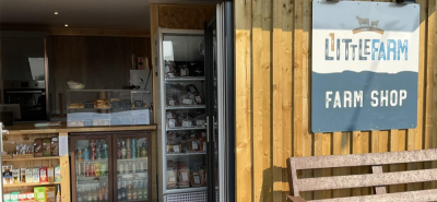 Store counter displays various goods; behind glass are baked items, below are beverages. The setting is a wooden farm shop with a sign reading “LITTLE FARM FARM SHOP” near a bench outside.