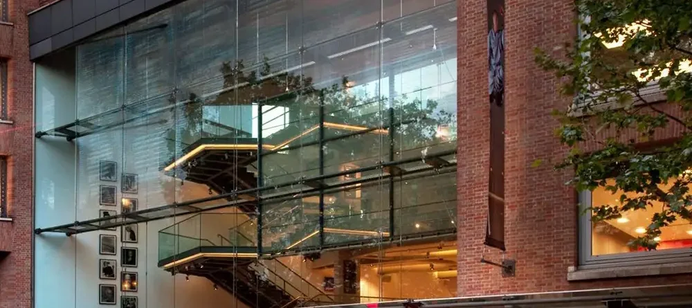 A modern glass staircase ascends inside a multi-story building, surrounded by framed pictures; the building's exterior features large glass windows and brick walls with nearby trees.