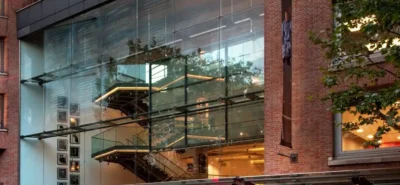 A modern glass staircase ascends inside a multi-story building, surrounded by framed pictures; the building's exterior features large glass windows and brick walls with nearby trees.