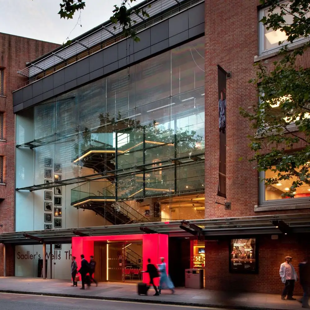 A modern glass staircase ascends inside a multi-story building, surrounded by framed pictures; the building's exterior features large glass windows and brick walls with nearby trees.