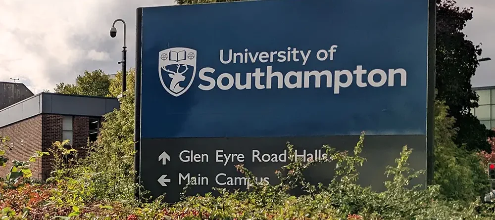 A blue sign reads "University of Southampton" with directional arrows pointing to Glen Eyre Road Halls and Main Campus, surrounded by green shrubs, against a backdrop of buildings and trees.