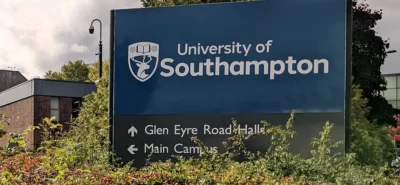 A blue sign reads "University of Southampton" with directional arrows pointing to Glen Eyre Road Halls and Main Campus, surrounded by green shrubs, against a backdrop of buildings and trees.