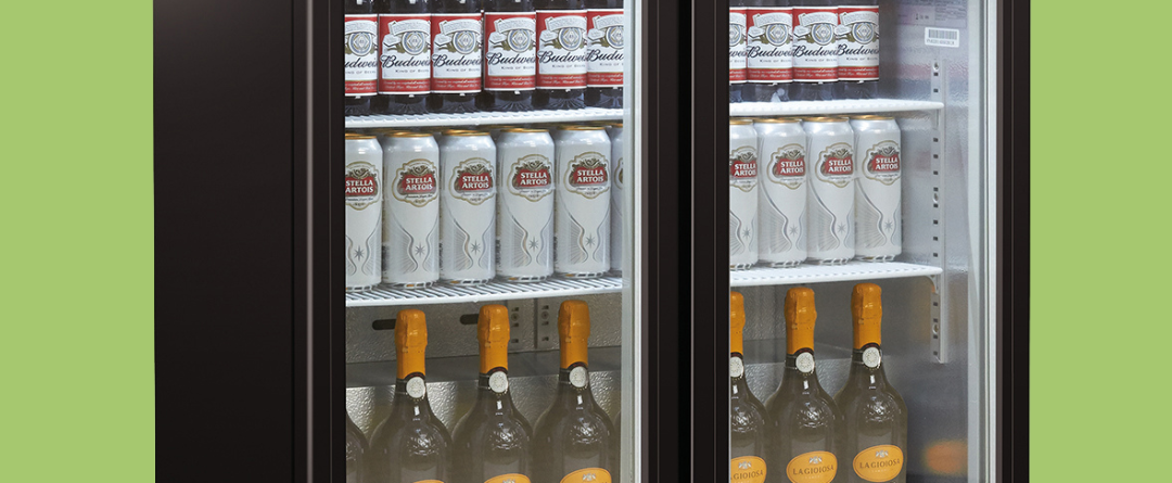 A refrigerated display case contains stacked Budweiser bottles, Stella Artois cans, and La Gioiosa orange-capped bottles, organized on three shelves in a retail environment.