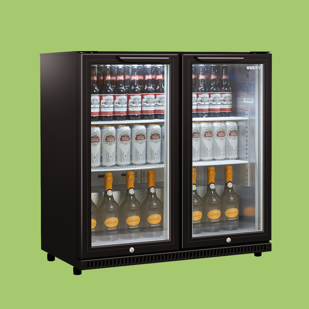 A refrigerated display case contains stacked Budweiser bottles, Stella Artois cans, and La Gioiosa orange-capped bottles, organized on three shelves in a retail environment.