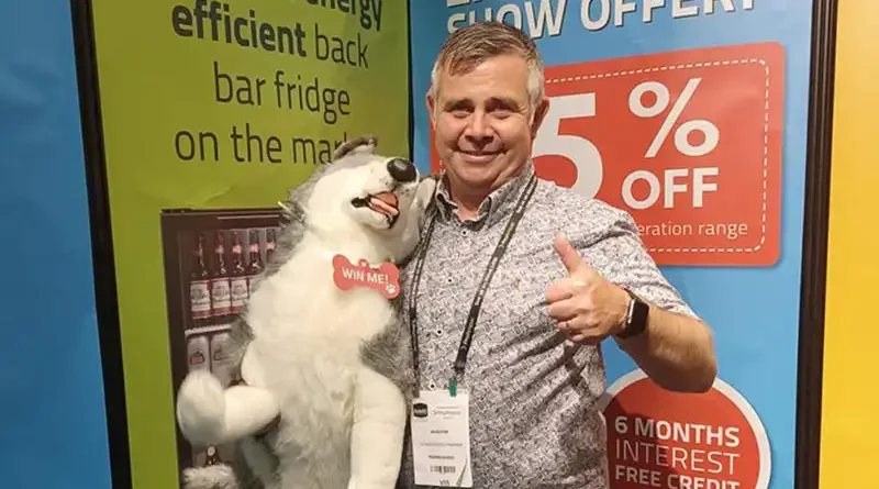 A smiling man, holding a plush husky toy, gives a thumbs up in front of colorful promotional signage displaying "WIN ME!" on the toy and "25% OFF" alongside details of interest-free credit.