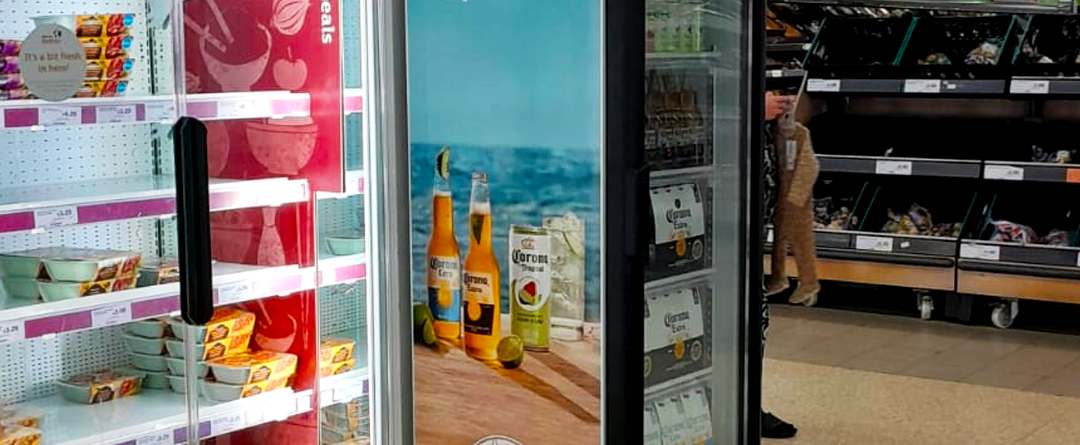 A refrigerated display case holds packaged food items beside a glass-fronted cooler advertising Corona beer with an ocean scene. A person stands near a produce section in a grocery store.