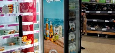 A refrigerated display case holds packaged food items beside a glass-fronted cooler advertising Corona beer with an ocean scene. A person stands near a produce section in a grocery store.