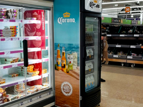 A refrigerated display case holds packaged food items beside a glass-fronted cooler advertising Corona beer with an ocean scene. A person stands near a produce section in a grocery store.