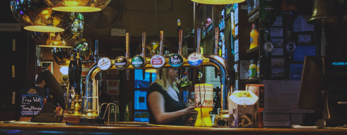 A classic British pub bar with beer taps, warm lighting, and a bartender serving customers, creating an inviting atmosphere for socialising and enjoying draft beers.