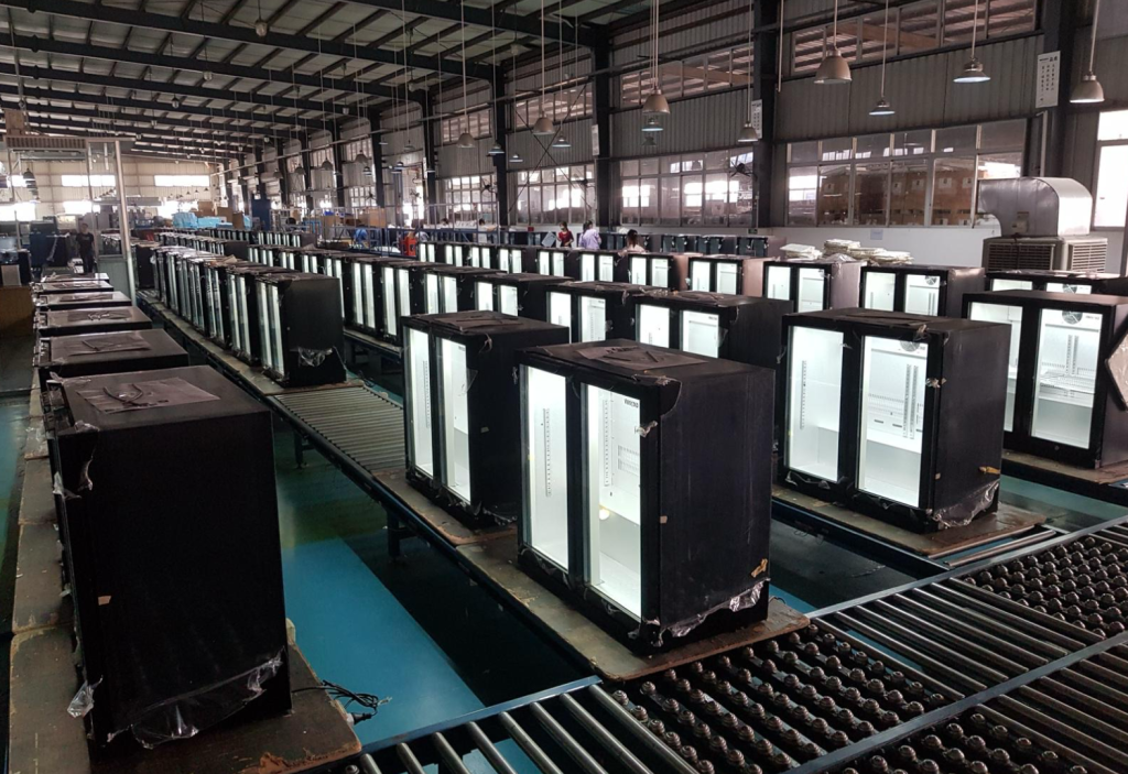 Husky refrigeration manufacturing facility showcasing a production line of glass-door commercial fridges in various stages of assembly. The well-lit factory floor highlights Husky’s large-scale production capabilities and commitment to quality in commercial refrigeration