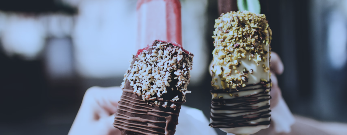 Close-up of hands holding gourmet ice lollies covered in chocolate and nuts, with a blurred outdoor background.