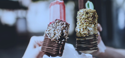 Close-up of hands holding gourmet ice lollies covered in chocolate and nuts, with a blurred outdoor background.
