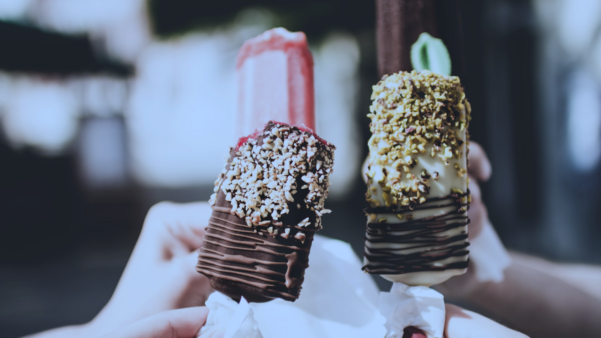 Close-up of hands holding gourmet ice lollies covered in chocolate and nuts, with a blurred outdoor background.